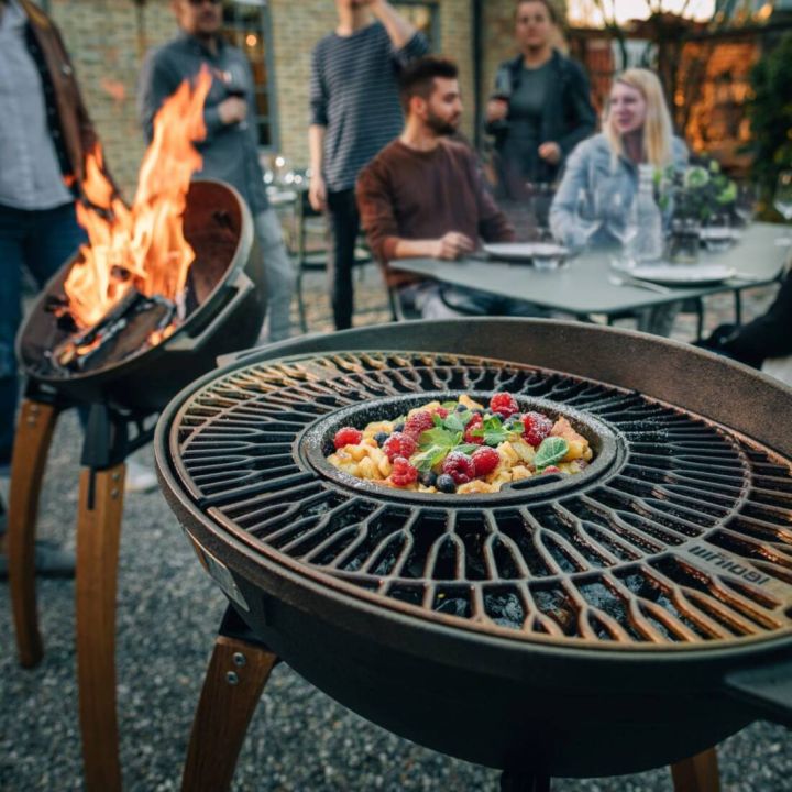 Eine Grillpfanne auf einem Outdoor-Grill, gefüllt mit Früchten und Minzblättern. Im Hintergrund sitzen mehrere Personen an einem Tisch, teils mit Getränken, während ein zweiter Grill mit Flammen zu sehen ist.