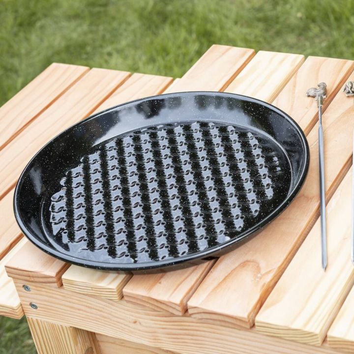 Schwarze, runde Grillpfanne mit Noppenstruktur, liegt auf einem hellen Holztisch im Freien; rechts daneben ein langer Metallspieß mit Haken am oberen Ende.